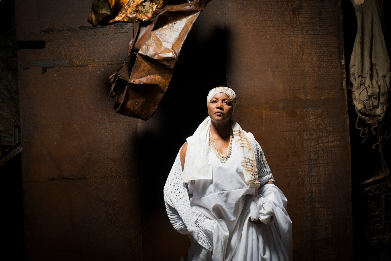 Mulher negra com vestido branco e dourado sentada olhando fixamente para a frente. No fundo da imagem, há um papelão com a cor marrom e um carrinho de mão pendurado.